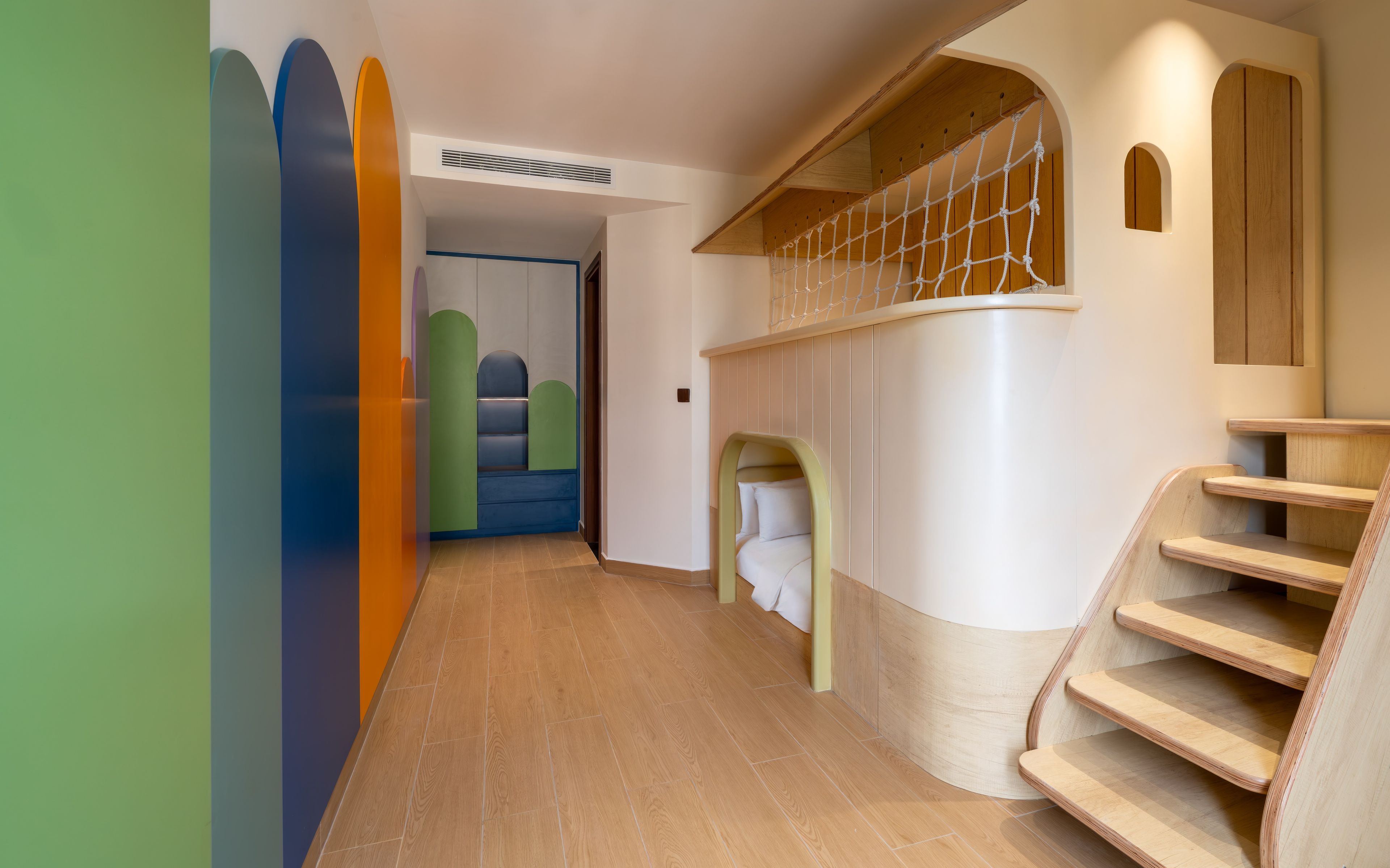 Un interior moderno con una escalera de madera, un muro curvo con diseño de red y un pasillo con paneles de pared geométricos y coloridos.