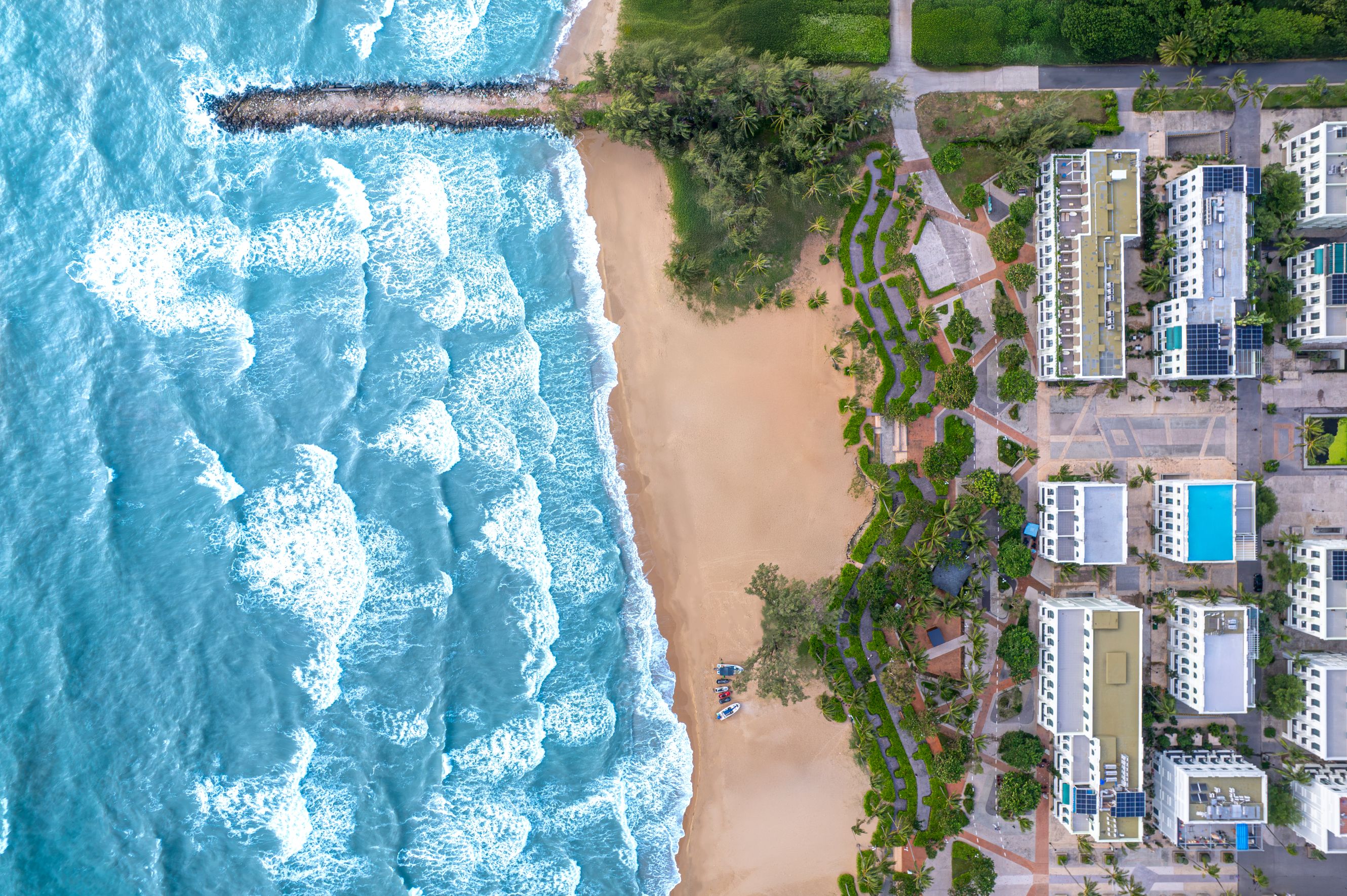 An aerial view of a coastal area with a sandy beach, a rocky breakwater, and a residential complex with multiple buildings and a swimming pool.