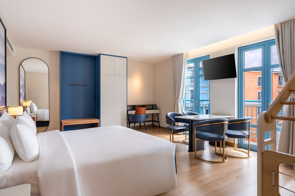 Ein Hotelzimmer mit Bett, Schreibtisch, zwei Stühlen, Fernseher und Blick auf die Stadt.