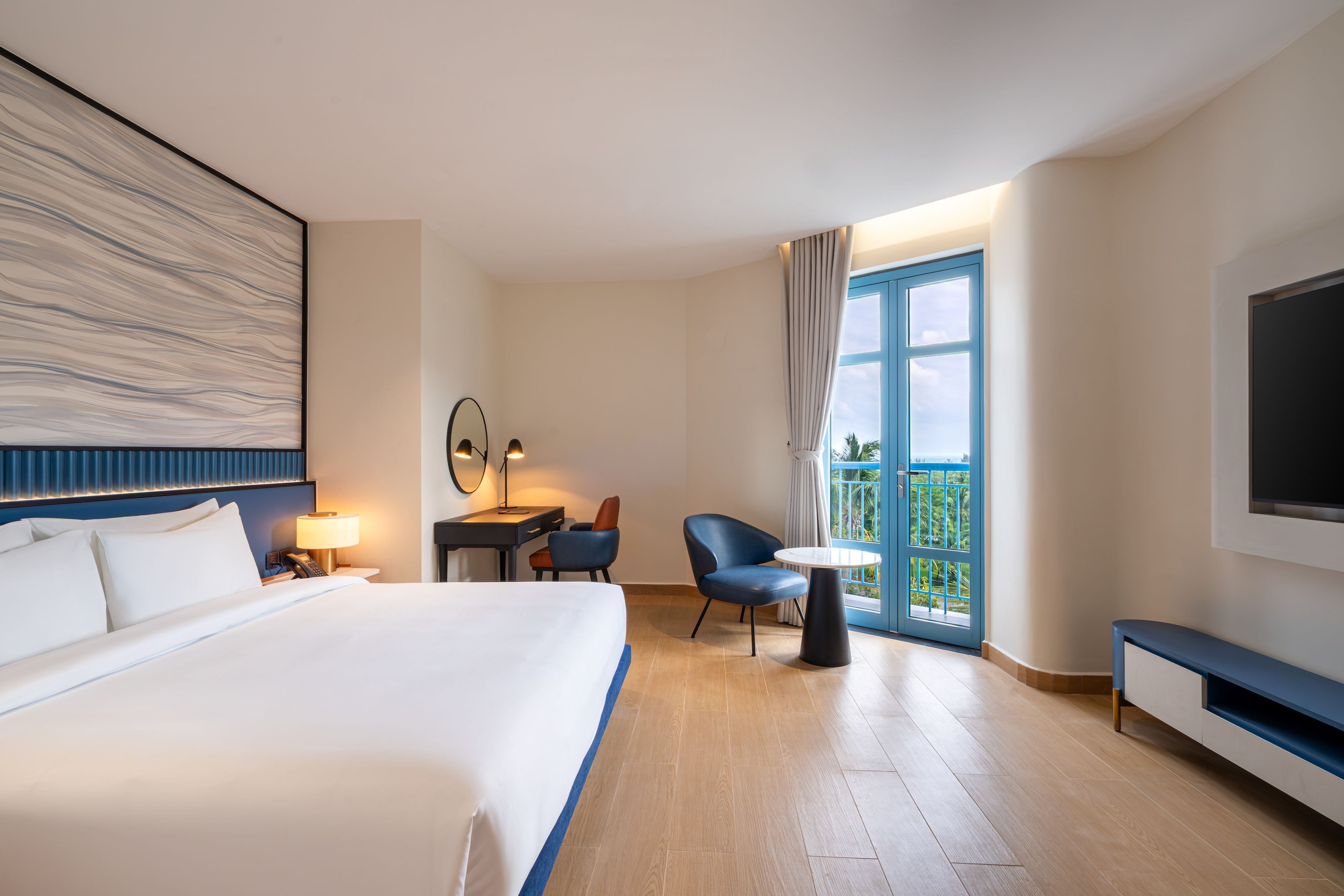 A hotel room with a large bed, a desk with a chair, a small table, a TV, and a balcony with a view.