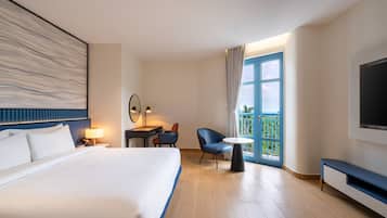 Quarto de hotel com uma cama grande, uma escrivaninha com cadeira, uma mesa pequena, televisão e varanda com vista.