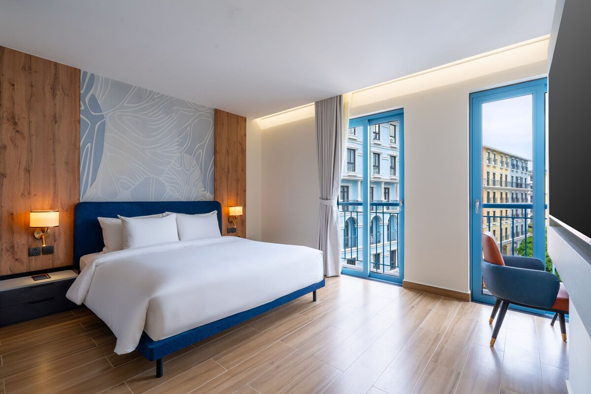 Ein modernes Hotelzimmer mit einem großen Bett, Blick auf die Stadt durch einen Balkon und einem blauen Sessel.