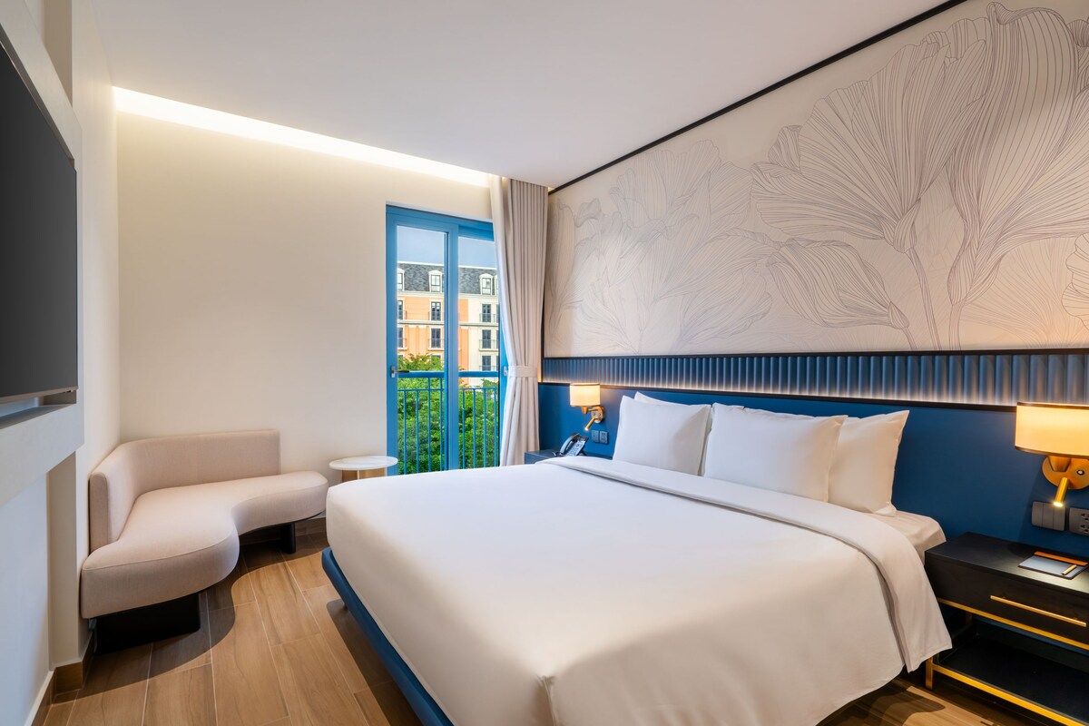Habitación de hotel con una cama grande, un escritorio con lámpara, una silla y vista a edificios por la ventana.