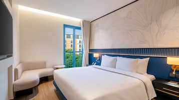 Ein Hotelzimmer mit einem großen Bett, einer Sitzecke, einem Schreibtisch und Blick auf Gebäude durch ein Fenster.