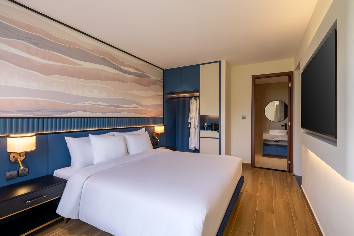 Ein modernes Hotelzimmer mit einem großen Bett, Nachttischlampen, einem Kleiderschrank, einem Spiegel und einem Badezimmer mit Waschbecken und Toilette.