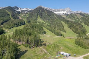 A mountainous landscape with a small building and a bus, surrounded by dense forests and snow-capped peaks.