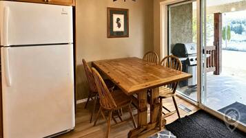 Une cuisine équipée d’un réfrigérateur, de meubles de rangement en bois, d’une table à manger avec des chaises, d’un lustre et d’une porte-fenêtre coulissante donnant sur un espace extérieur.