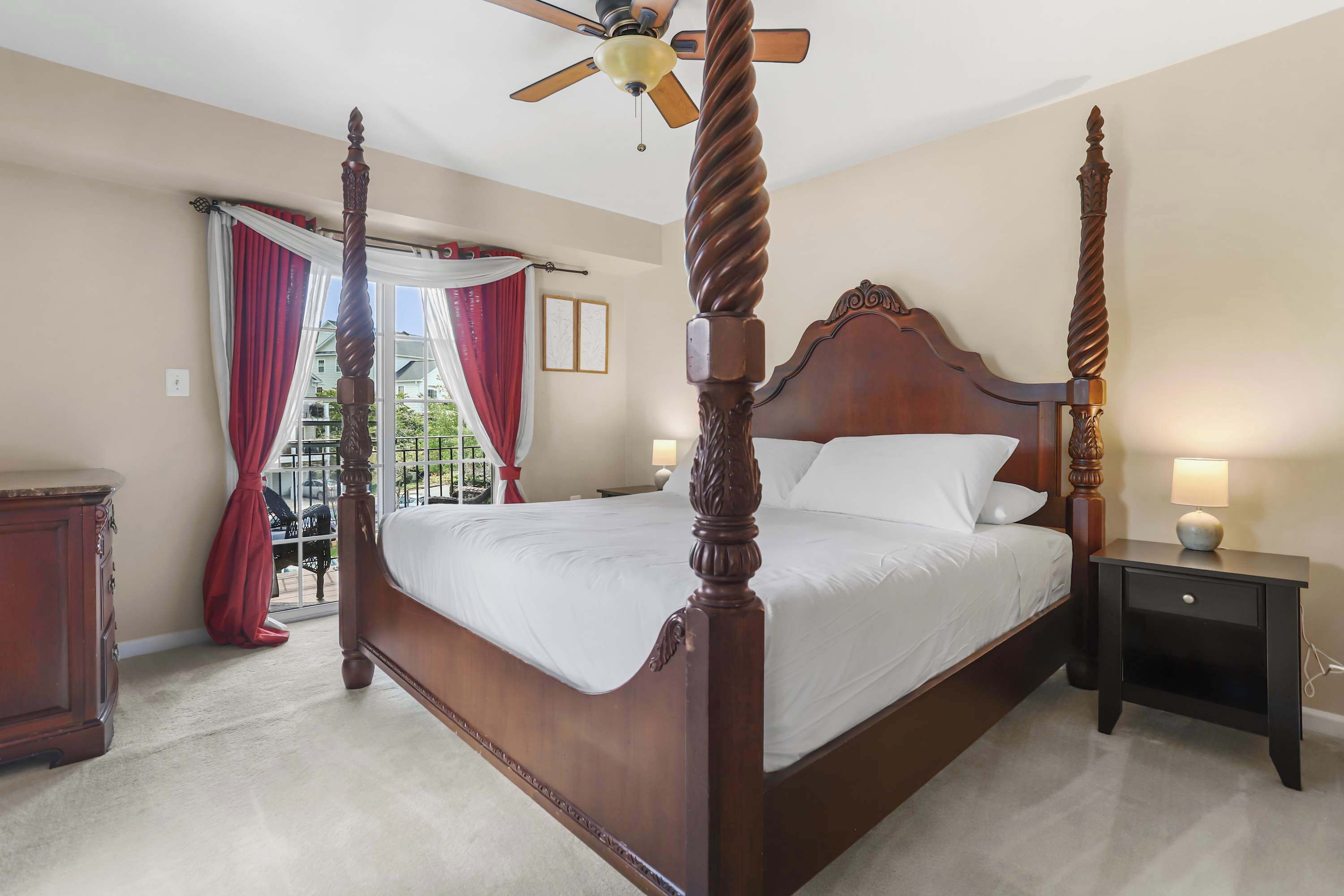 A four-poster bed with a wooden headboard and footboard, a bedside table with a lamp, and a view of a balcony through a window with red curtains.