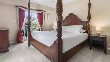 A four-poster bed with a wooden headboard and footboard, a bedside table with a lamp, and a view of a balcony through a window with red curtains.