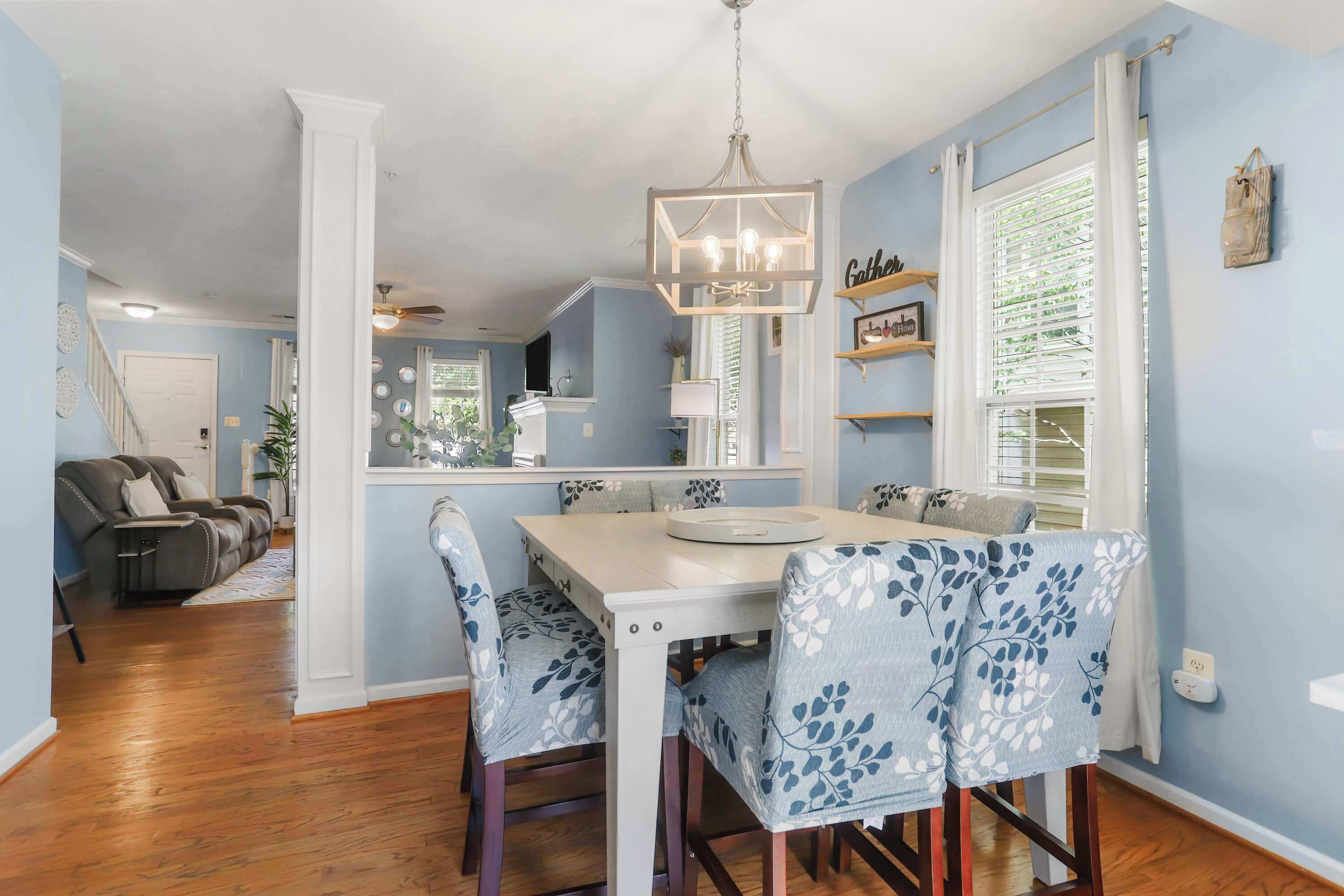 A dining area with a white table and blue floral chairs, a chandelier, and a view of a living room with a sofa and a ceiling fan.