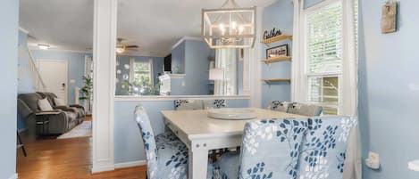A dining area with a white table and blue floral chairs, a chandelier, and a view of a living room with a sofa and a ceiling fan.