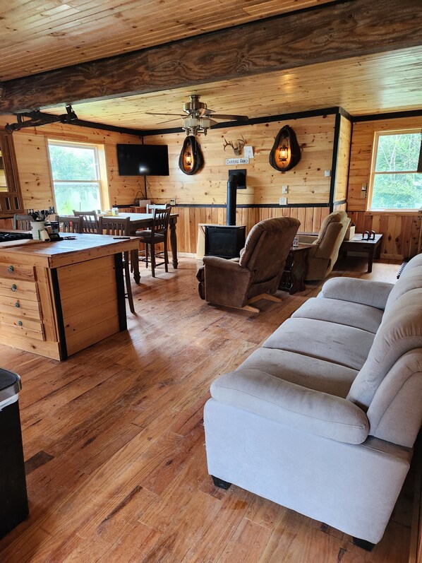A cabin interior with a wooden floor, a sofa, a dining table, chairs, a fireplace, and a ceiling fan. - Hunter's Paradise in Northeast Montana (Culbertson)