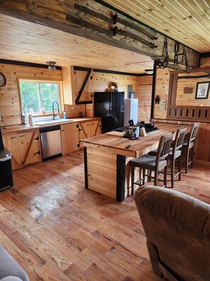 A rustic cabin interior with a wooden floor, a kitchen area with a black refrigerator, a dining table with chairs, and a wooden ceiling with exposed beams.