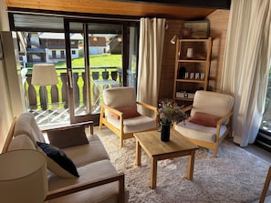 Habitació acollidora amb un sofà, dues butaques, una taula de centre i vista a un balcó amb moble d'exterior.