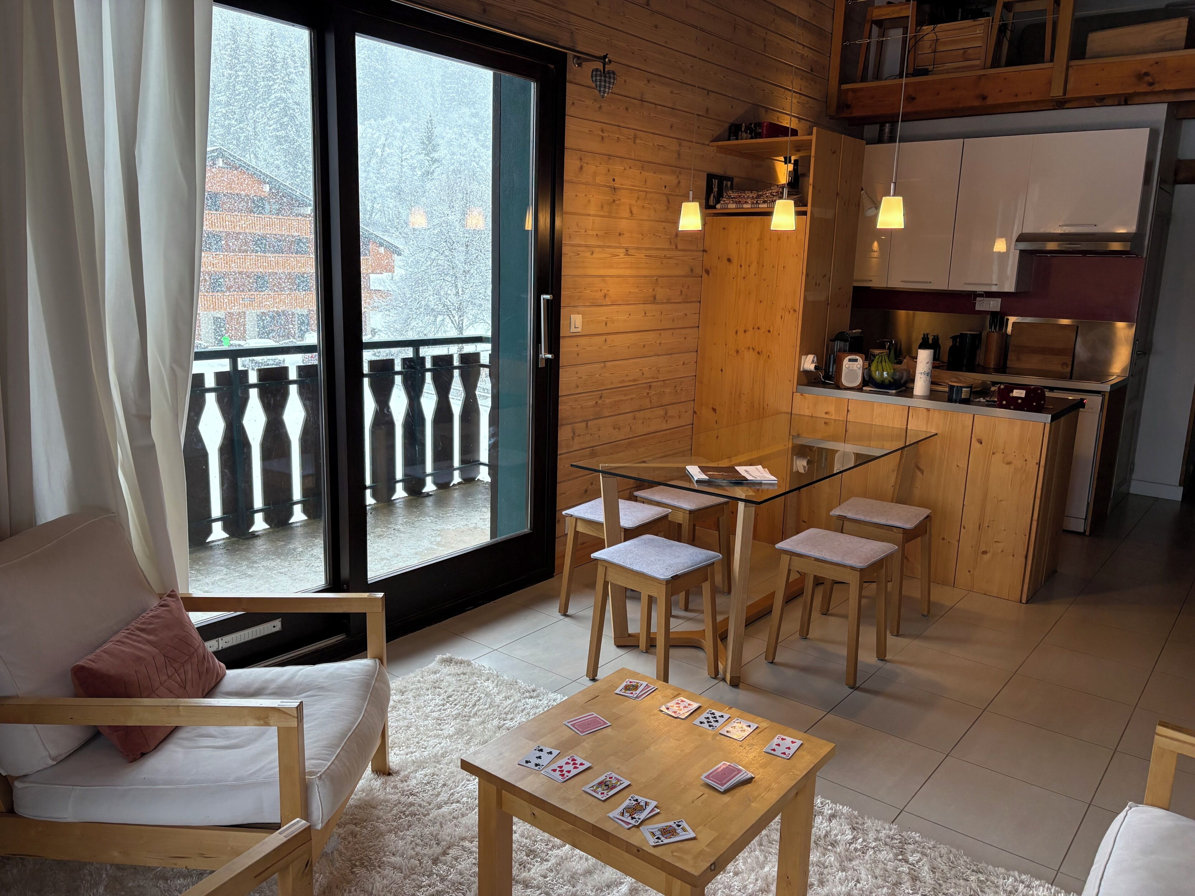 A cozy room with a sofa, a glass-top table with cards, a wooden kitchen, and a balcony with a view of snow-covered buildings.