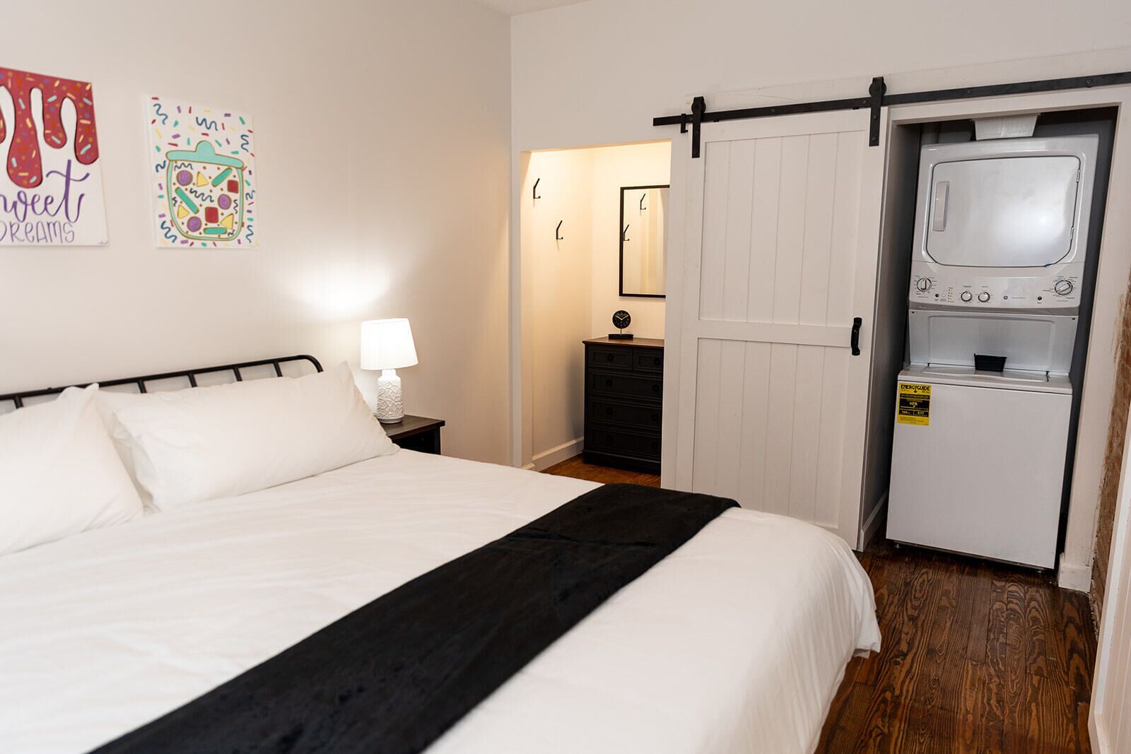 A bedroom with a bed, a nightstand, a lamp, a dresser, a mirror, and a sliding door leading to a laundry area with a washing machine.