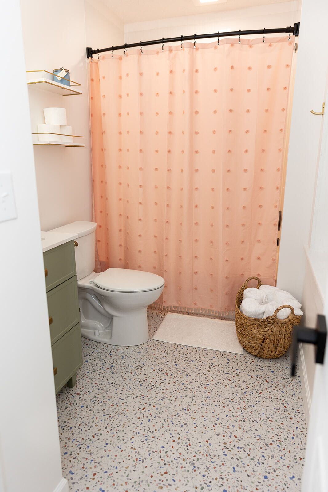 A bathroom with a toilet, a wicker basket with towels, a shower curtain, and a speckled floor.