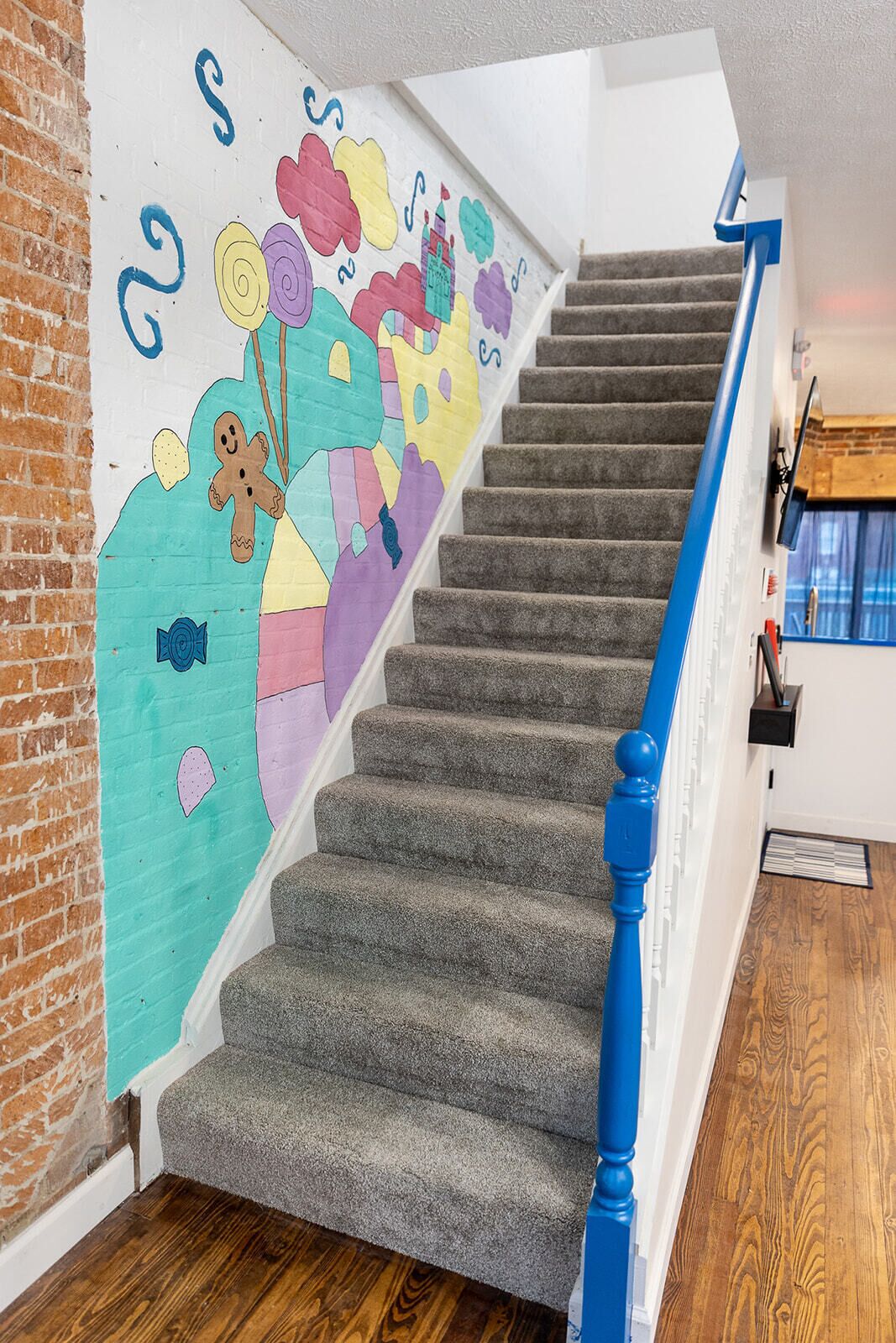 Una escalera con barandilla azul, peldaños alfombrados y un mural en la pared con diversas formas y diseños coloridos.