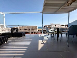 Een overdekt terras met uitzicht op zee, met een bank, stoelen en een tafel.