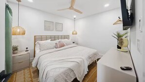 Quarto com cama, mesa de cabeceira, ventilador na parede e televisão sobre um suporte.
