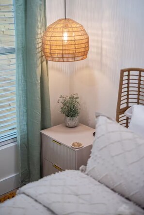 Ein Schlafzimmer mit einem Bett, einem Nachttisch mit Lampe, einer Zimmerpflanze und einem Fenster mit Vorhängen.