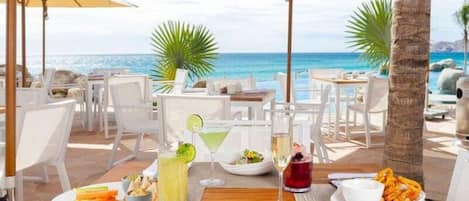 An outdoor dining area with tables, chairs, and umbrellas overlooking a beach.