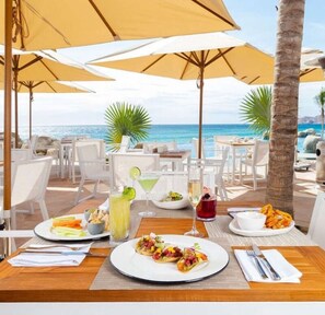 Un área de comedor al aire libre con mesas, sillas y sombrillas con vista a una playa.