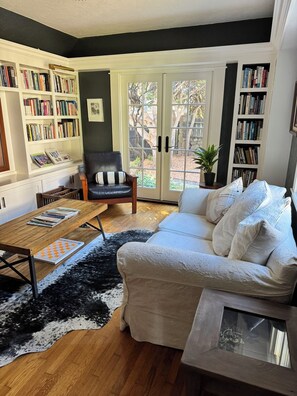 Ein Wohnzimmer mit einem Holztisch, einem schwarzen Ledersessel, einem weißen Sofa und einem schwarz-weißen Teppich. An beiden Seiten des Raumes befinden sich Bücherregale und ein großes Fenster mit Blick auf Bäume.