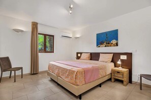 A bedroom with a bed, a nightstand, a chair, a window with curtains, an air conditioning unit, and a wall-mounted painting. - Bon Bida Buriku with shared pool, Hato (Kralendijk)
