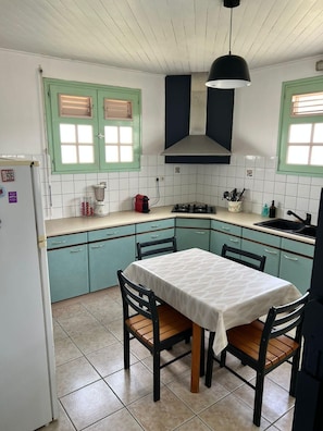 A kitchen with a dining table, chairs, a refrigerator, a stove, and a window with blinds. - Bouliki House (Saint-Joseph)