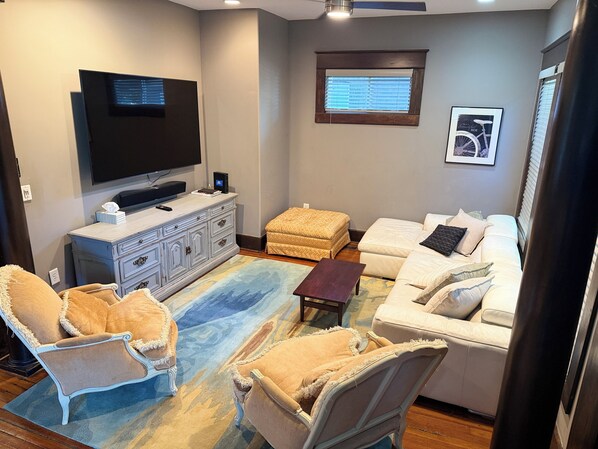 A living room with a television, a sofa, two armchairs, a coffee table, and a window with blinds.