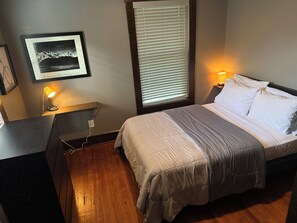 Un dormitorio con cama, lámparas de noche, mesita de noche, una fotografía enmarcada en la pared y una ventana con persianas.