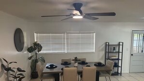Uma sala de jantar com uma mesa posta para quatro pessoas, um ventilador de teto e uma janela com persianas.