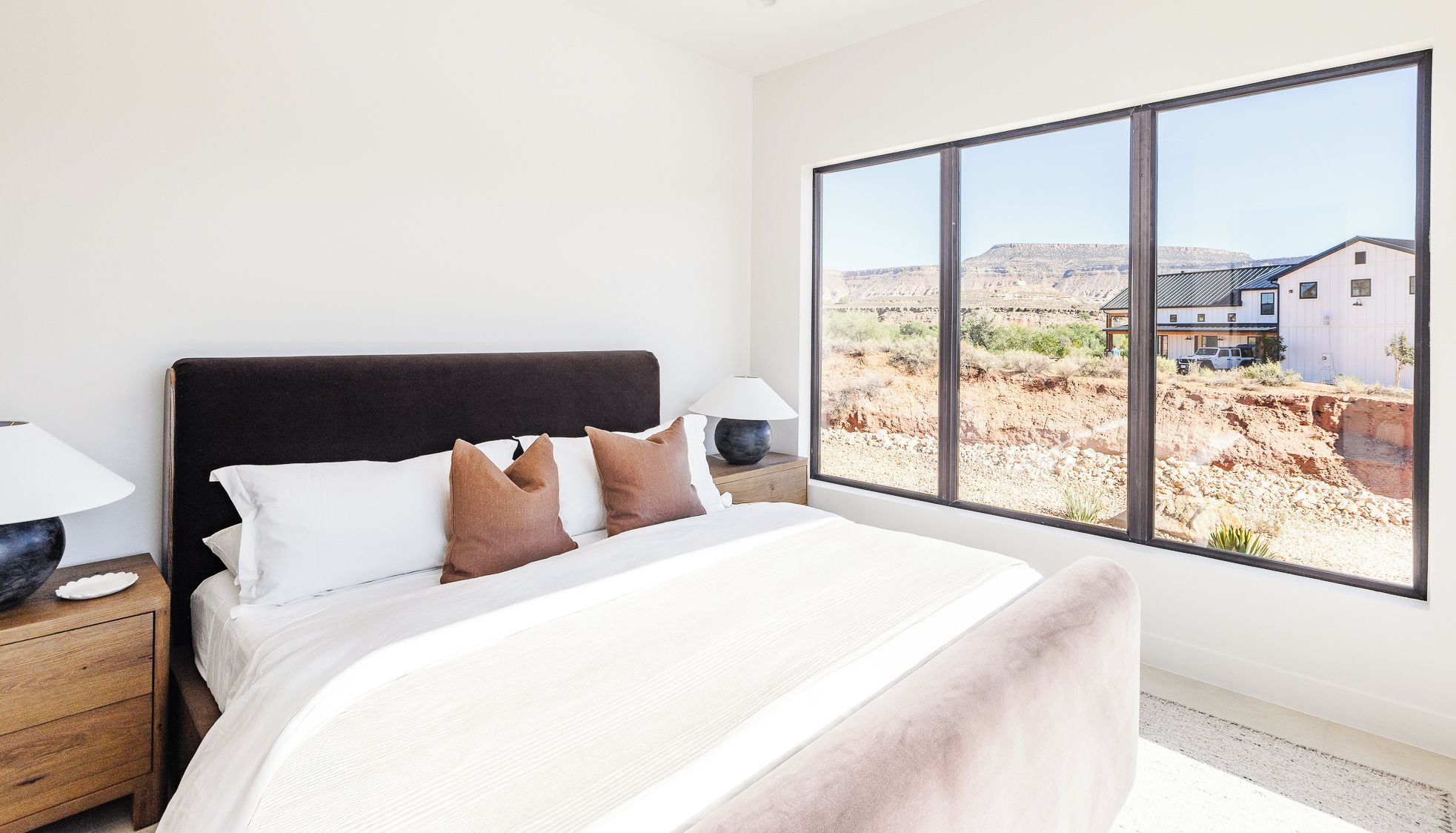 Un dormitorio con un ventanal grande, una cama con ropa blanca y almohadas marrones, una mesita de noche con lámpara, y vistas a un edificio y un paisaje.