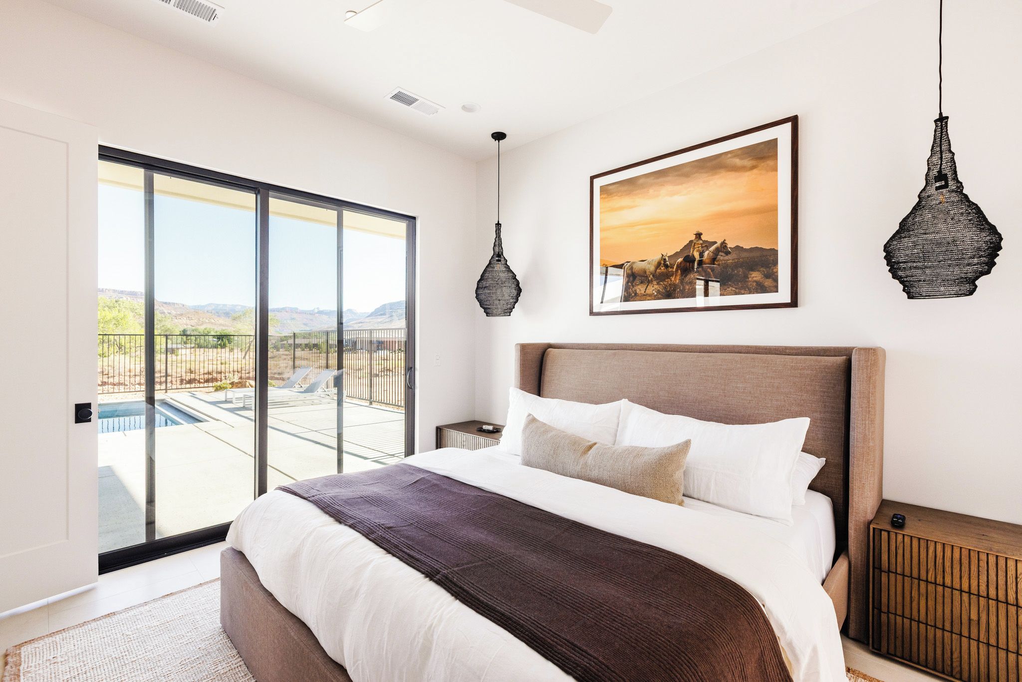 A bedroom with a large bed, a view of the outdoors through a sliding door, a framed picture on the wall, and a hanging light fixture.