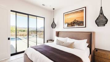 A bedroom with a large bed, a view of the outdoors through a sliding door, a framed picture on the wall, and a hanging light fixture.