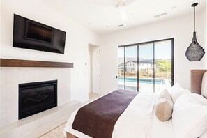Un dormitori amb una xemeneia, un llit amb almoades, un televisor fixat a la paret i vistes a una piscina i cases a l'exterior.