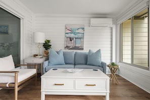 A room with a white sofa, a side table with a lamp, a painting, and a window with blinds. - A Place to Remember | Seabrook Convenience (Seabrook Island)