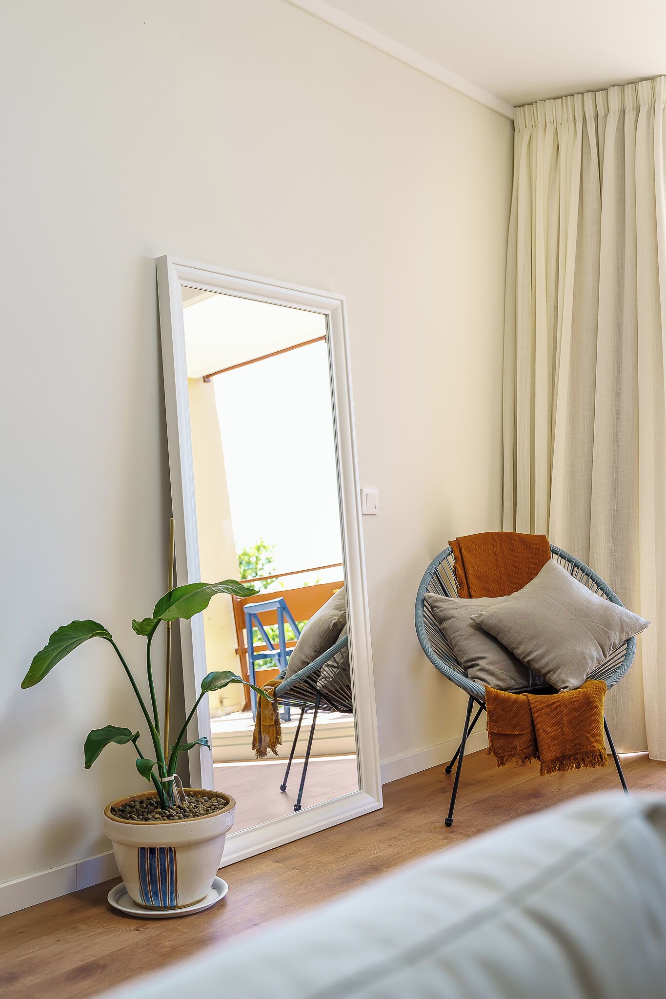 Quarto com um espelho grande, um vaso de planta, uma cadeira com almofadas e vista através de uma porta.