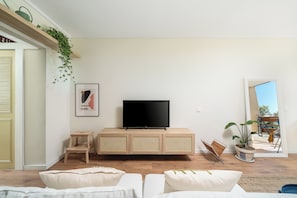 Ein Wohnzimmer mit Flachbildfernseher, einer Rattan-Konsole, einem kleinen Holzschemel und einem Sofa.