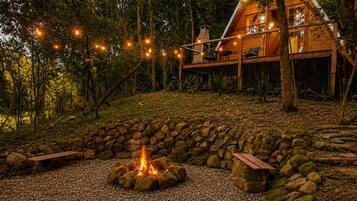 A wooden cabin with a stone fire pit surrounded by a gravel area, set in a forested environment.