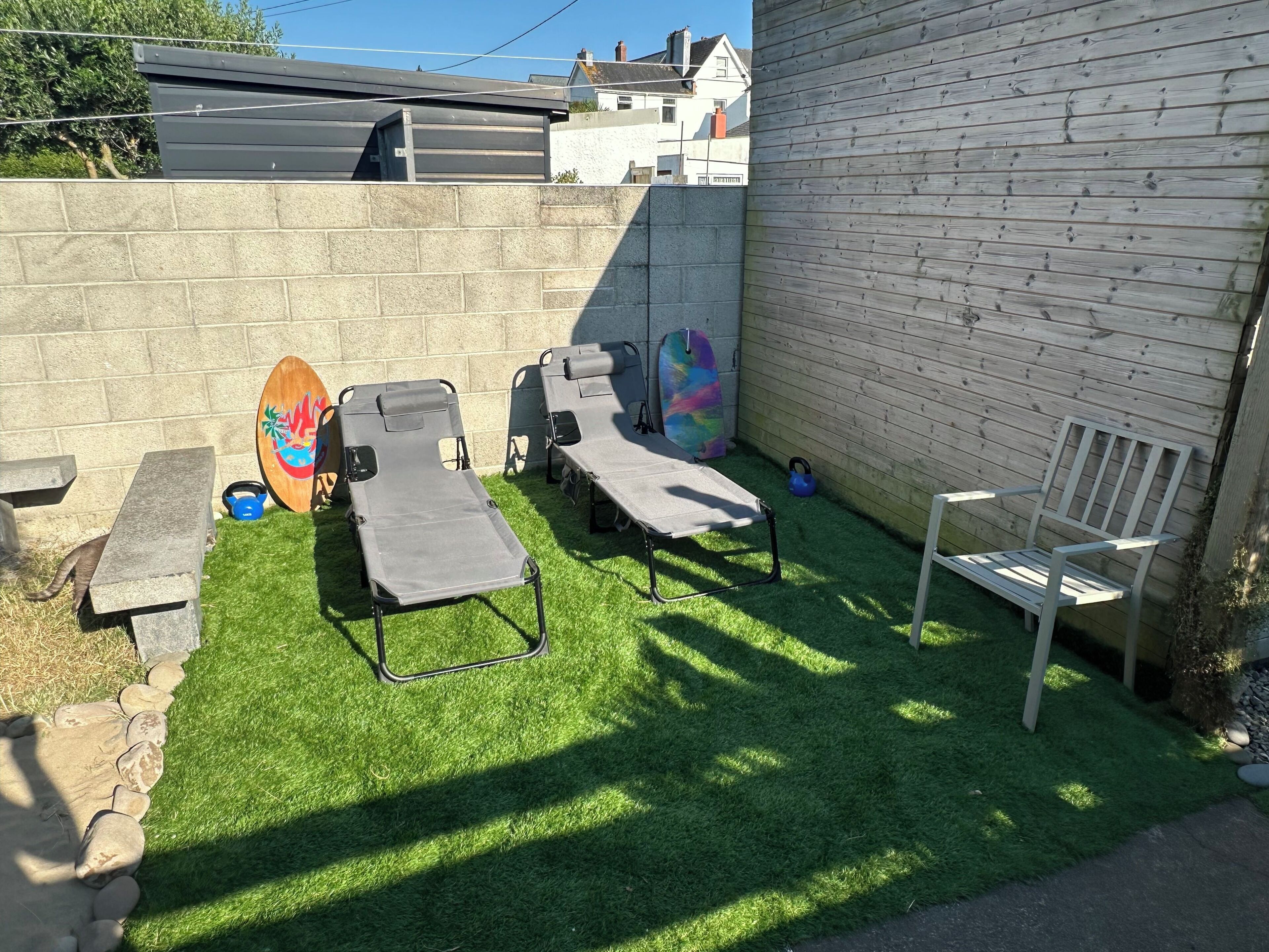 A backyard with artificial grass, two lounge chairs, a wooden surfboard, and a white bench.