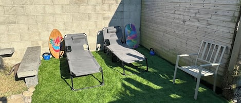 A backyard with artificial grass, two lounge chairs, a wooden surfboard, and a white bench.