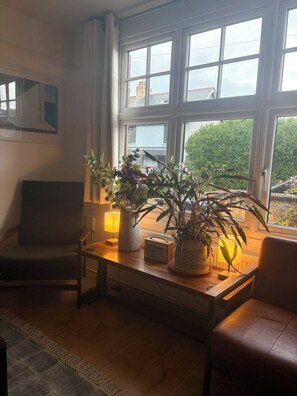 Ein Zimmer mit großem Fenster, einem Holztisch, einer Topfpflanze, einer Lampe und einem Stuhl.