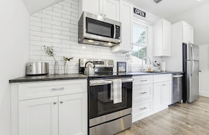 A modern kitchen with white cabinetry, stainless steel appliances, and a window with an 'OPEN' sign. - May River Cottage on Lawrence street, old Town. (Bluffton)