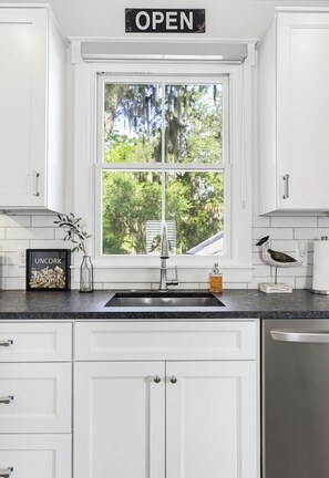 A kitchen with white cabinets, a black countertop, a stainless steel sink, and a window with a view of trees.