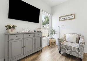 A room with a grey cabinet, a television, a patterned armchair, a window with a view, and a welcome sign. - May River Cottage on Lawrence street, old Town. (Bluffton)