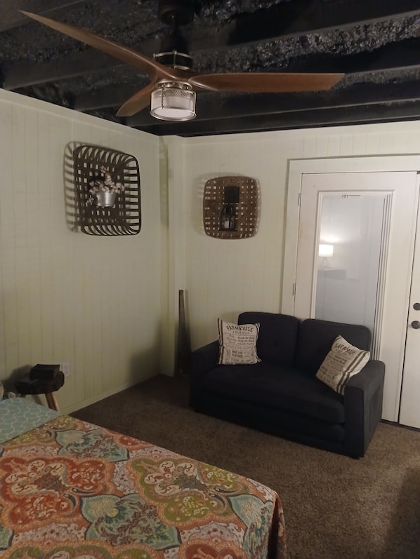 A room with a ceiling fan, a patterned bedspread, a dark-colored sofa with a pillow, and a white door.