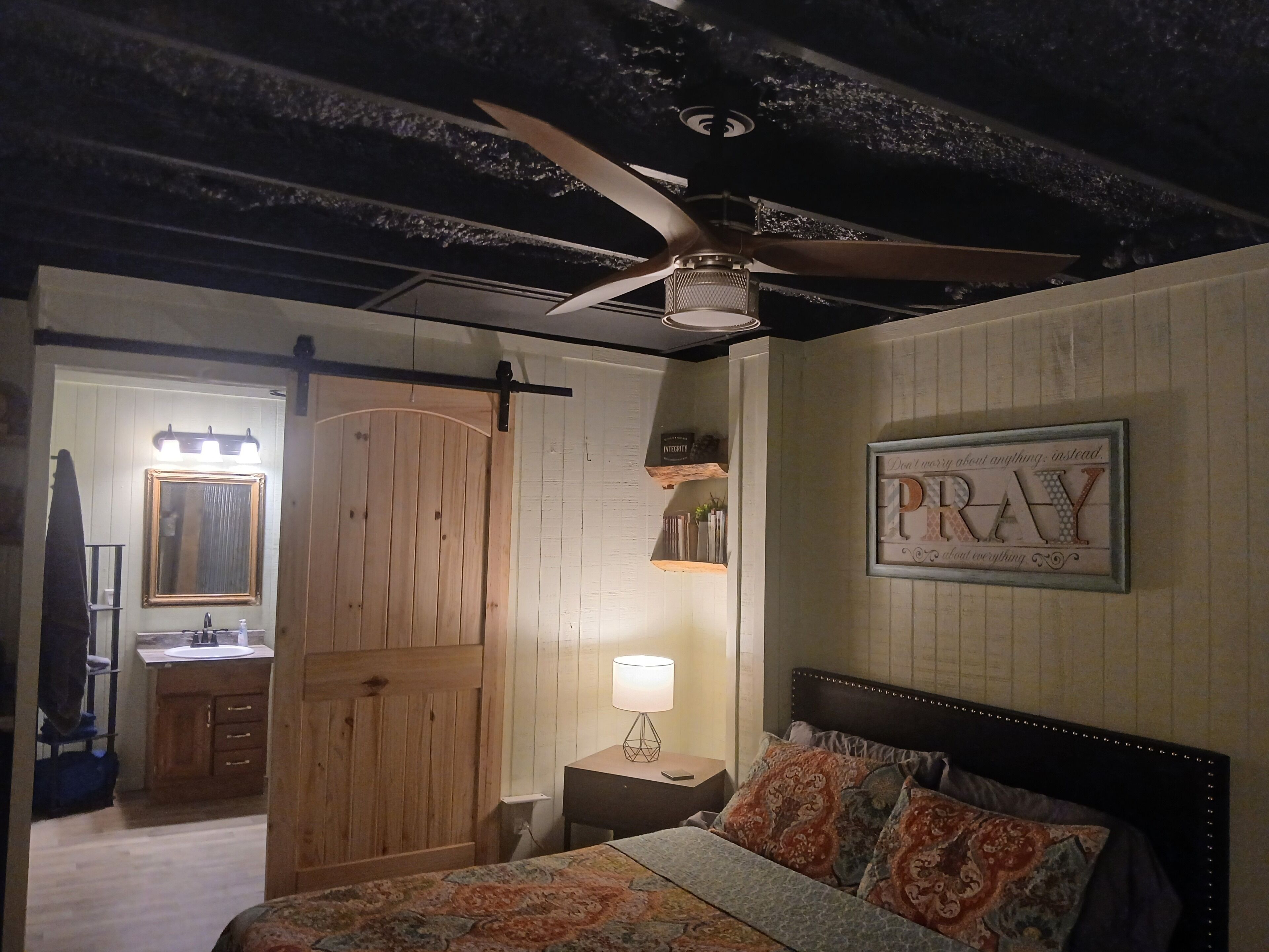 Ein Schlafzimmer mit einem Bett, einem Nachttisch, einer Lampe, einem Deckenventilator, einer Holztür und einem an der Wand befestigten Schild.