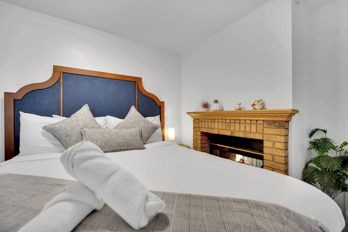 A neatly made bed with a headboard, a fireplace with decorative items, and a potted plant.